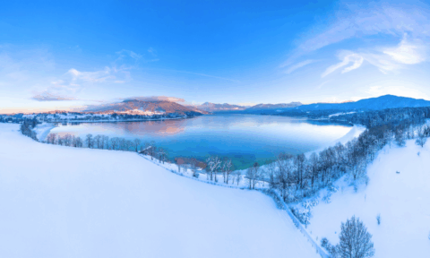 Winterpanorama am Tegernsee mit schneebedeckten Wiesen und Blick auf die umliegenden Alpen, klare blaue Himmel und spiegelndes Wasser – idyllische Landschaft nahe Bad Wiessee.
