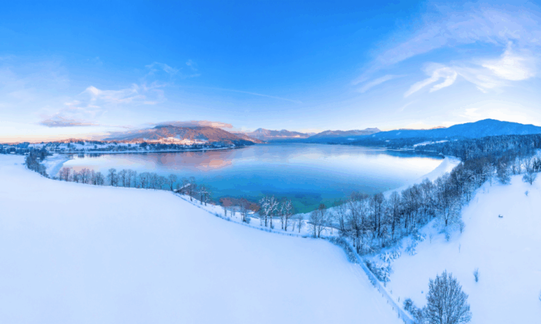 Winterpanorama am Tegernsee mit schneebedeckten Wiesen und Blick auf die umliegenden Alpen, klare blaue Himmel und spiegelndes Wasser – idyllische Landschaft nahe Bad Wiessee.